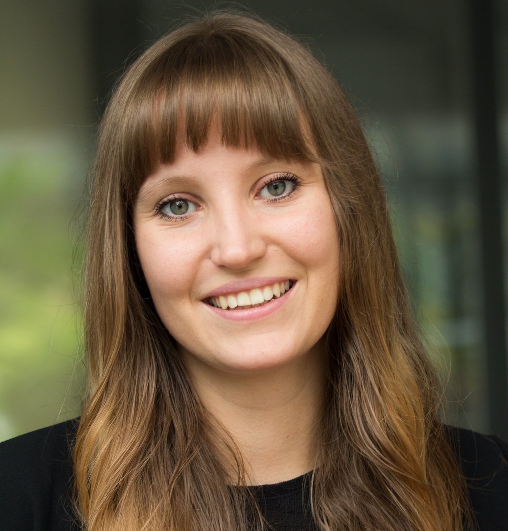 A young woman with long, wavy brown hair and bangs smiles warmly at the camera, wearing a black shirt. The background is softly blurred.