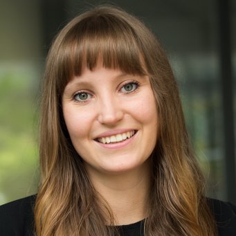 A young woman with long, wavy brown hair and bangs smiles warmly at the camera, wearing a black shirt. The background is softly blurred.
