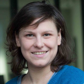 A close-up portrait of a smiling person with short, wavy brown hair, wearing a blue sweater.