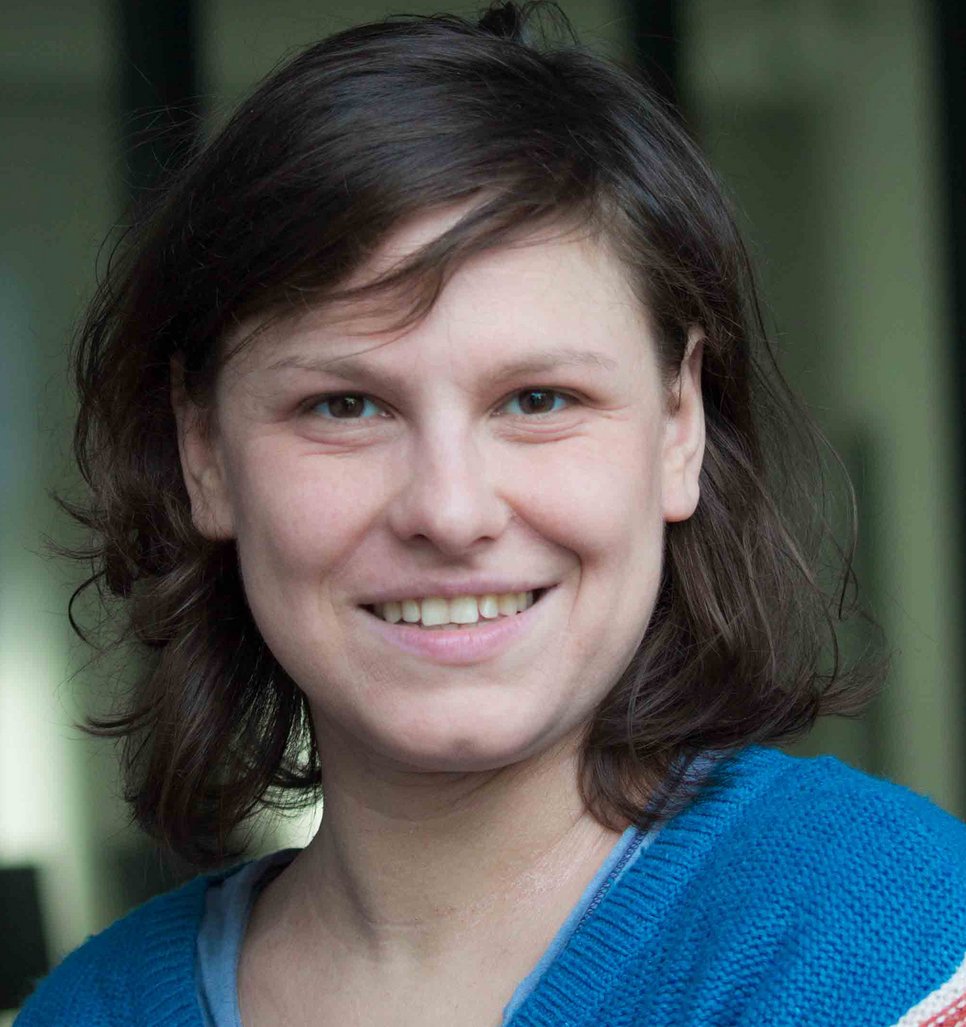 A close-up portrait of a smiling person with short, wavy brown hair, wearing a blue sweater.