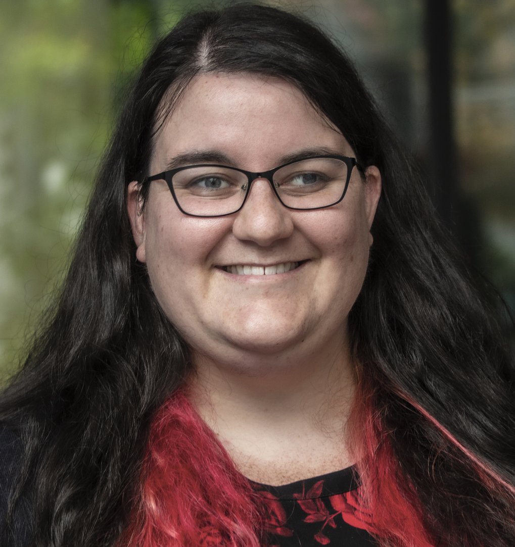A smiling person with long, dark hair featuring pink highlights, wearing glasses, and a black top.