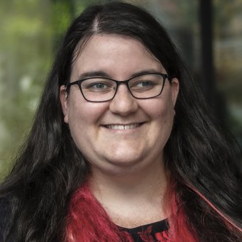 A smiling person with long, dark hair featuring pink highlights, wearing glasses, and a black top.