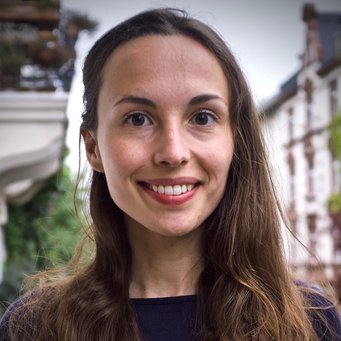 A woman with long, dark hair is smiling in an outdoor setting, with buildings in the background.