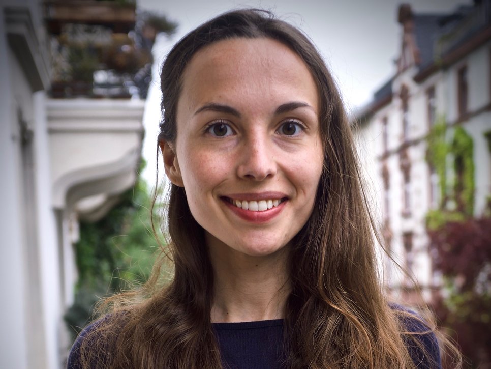 A woman with long, dark hair is smiling in an outdoor setting, with buildings in the background.