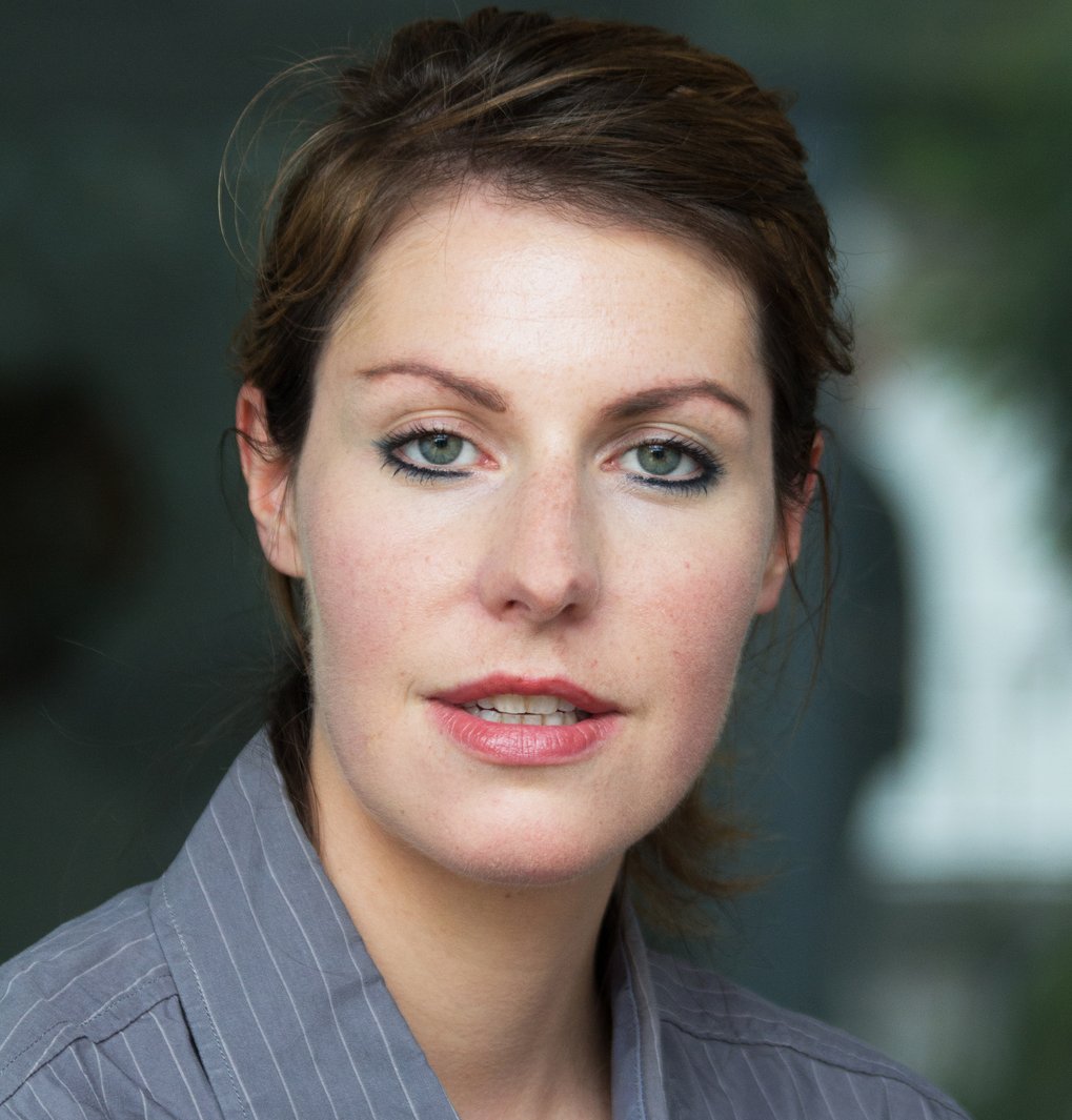 A woman with shoulder-length hair is looking at the camera, wearing a striped shirt. The background is blurred.