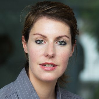 A woman with shoulder-length hair is looking at the camera, wearing a striped shirt. The background is blurred.
