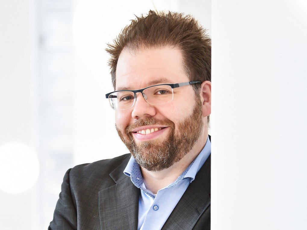 A smiling man with a beard, wearing glasses and a black blazer, against a light background.