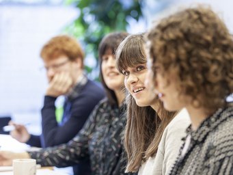 A group of four people engaged in conversation around a table, with two women smiling and looking at each other, and two men in the background.