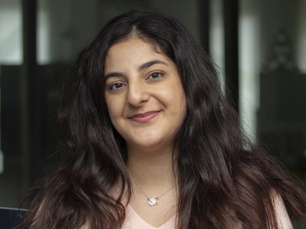 A smiling woman with long, wavy hair is sitting indoors. She wears a pink top and a necklace, with a blurred background of indoor settings.