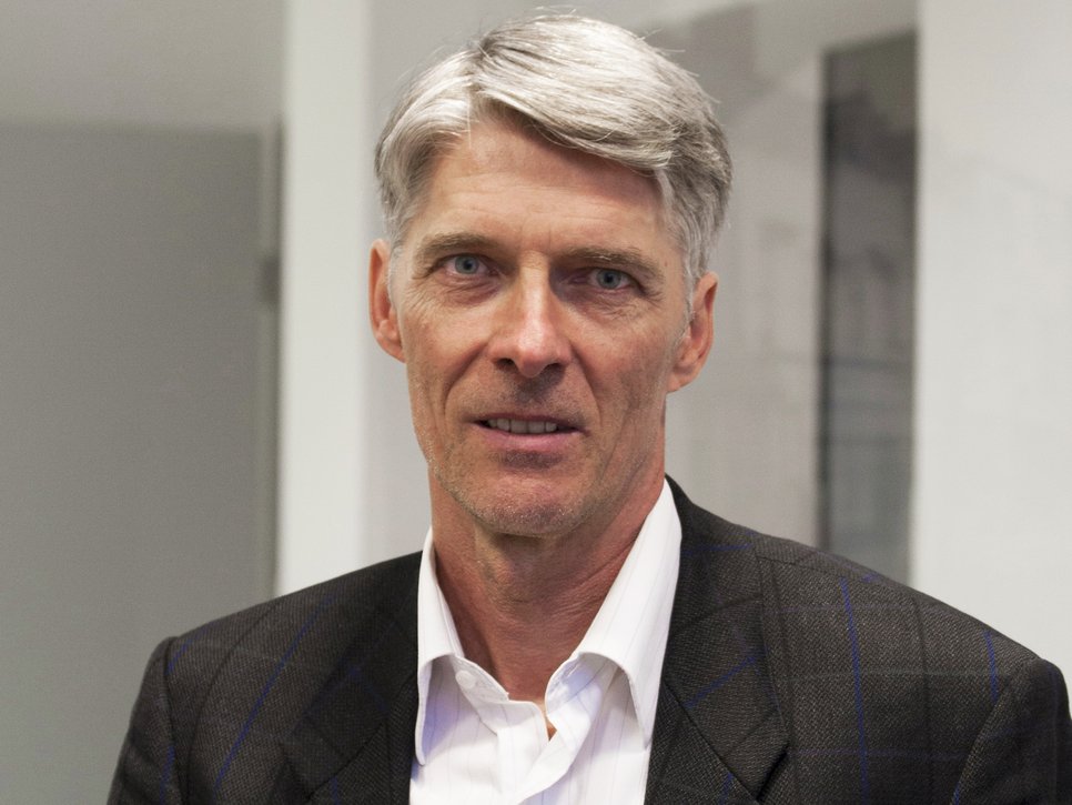 A man with gray hair, wearing a dark blazer over a white shirt, is looking directly at the camera.