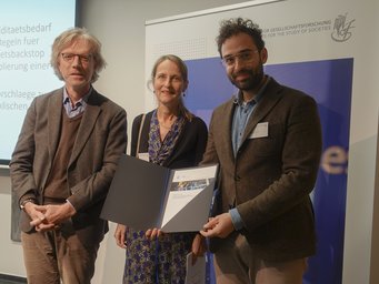 Three people stand together, holding a document. They are in front of a presentation backdrop. The setting appears to be a formal event or meeting.