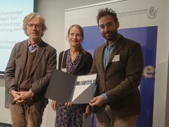 Three people stand together, holding a document. They are in front of a presentation backdrop. The setting appears to be a formal event or meeting.