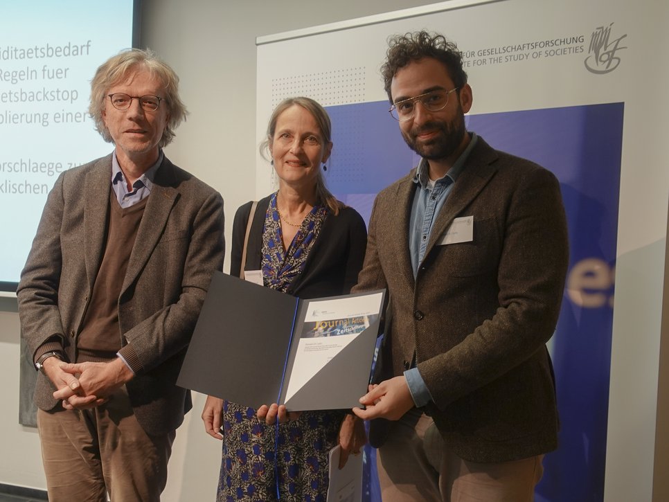 Three people stand together, holding a document. They are in front of a presentation backdrop. The setting appears to be a formal event or meeting.