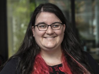 A woman with long black hair featuring pink highlights, wearing glasses and a black sweater, smiles at the camera.