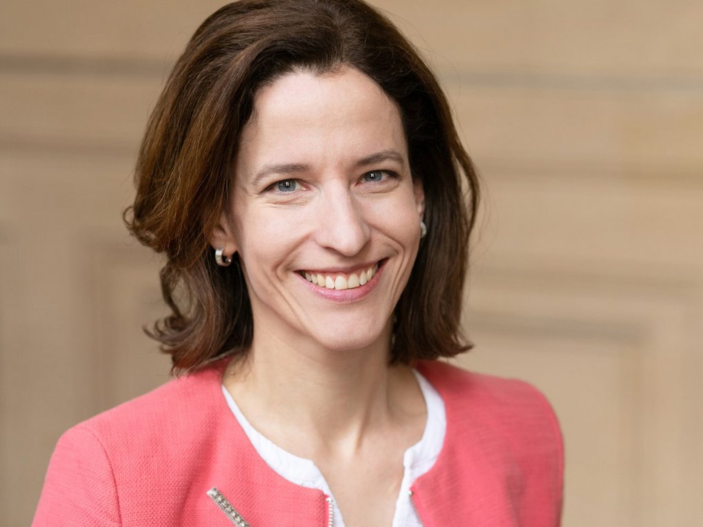 A woman with shoulder-length brown hair smiles, wearing a coral-colored blazer over a white top, with a neutral background.