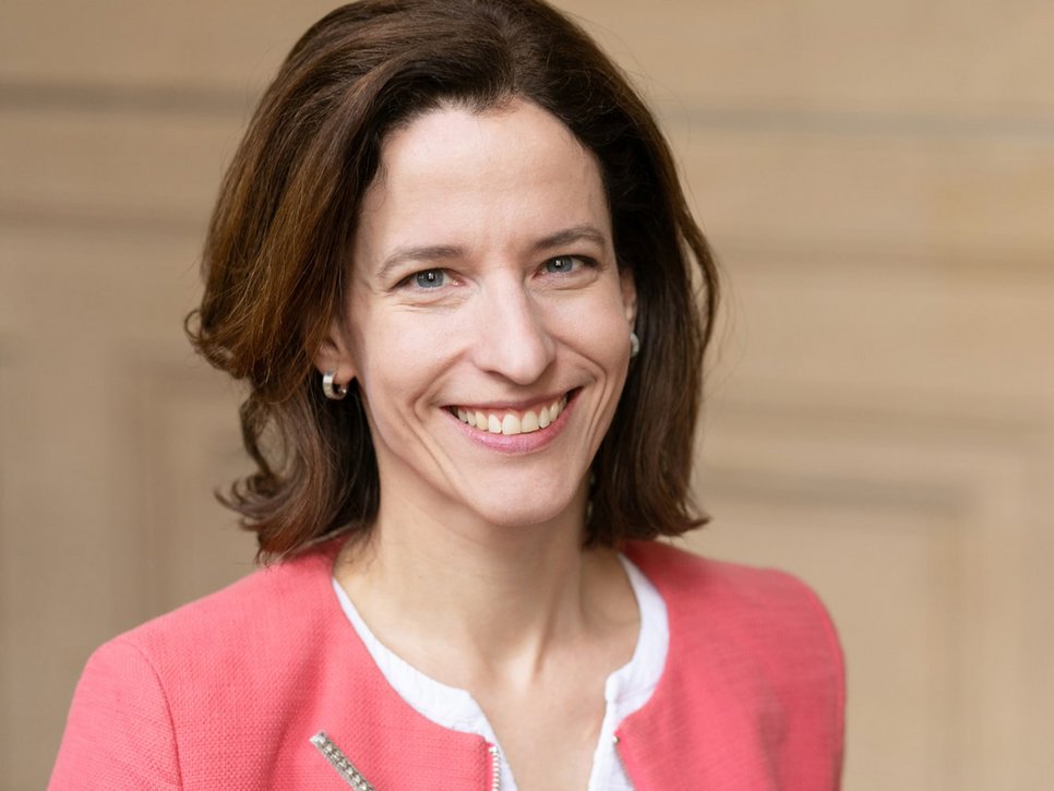 A woman with shoulder-length brown hair smiles, wearing a coral-colored blazer over a white top, with a neutral background.