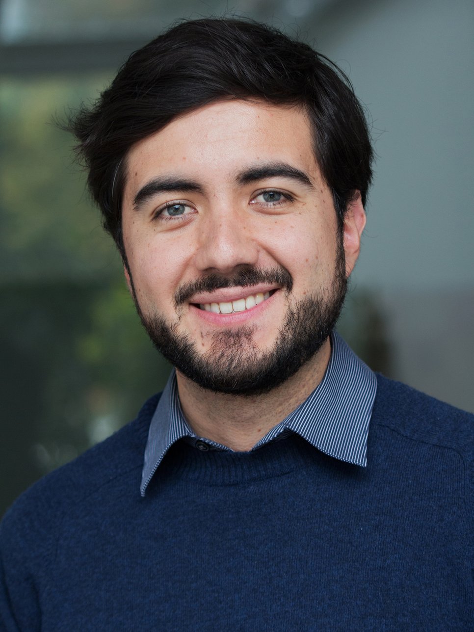 A smiling man with a beard, wearing a navy blue sweater and a collared shirt, is posing for the photo.