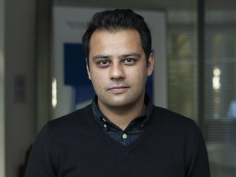 A man wearing a dark sweater and collared shirt is posing for a photo in an indoor setting.