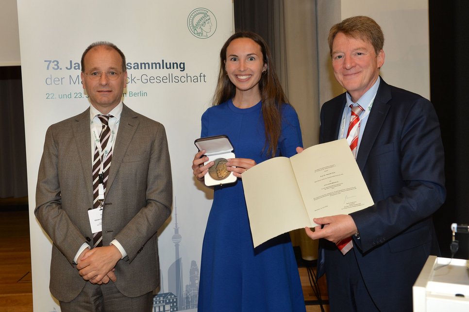 Three individuals at a formal event, with one woman in a blue dress holding a medal and a certificate, while the two men stand beside her. A banner in the background indicates a gathering of a society in Berlin.
