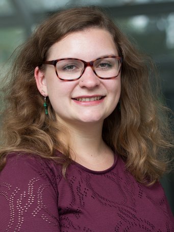 A smiling woman with curly brown hair, wearing glasses and green earrings, posing for a portrait in a casual purple top.