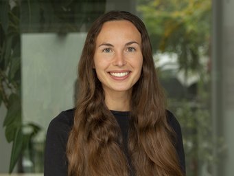 A smiling young woman with long, wavy hair, wearing a black top, stands in front of a blurred background with greenery.