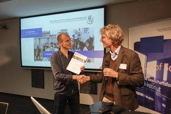 Two men are shaking hands at a formal event, one holding a document. A presentation screen in the background features images and text related to climate activism and the Max Planck Institute.