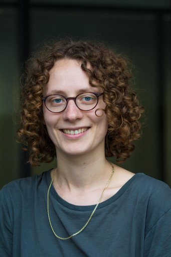 A smiling individual with curly hair, wearing glasses and a dark shirt, is photographed in front of a blurred background.