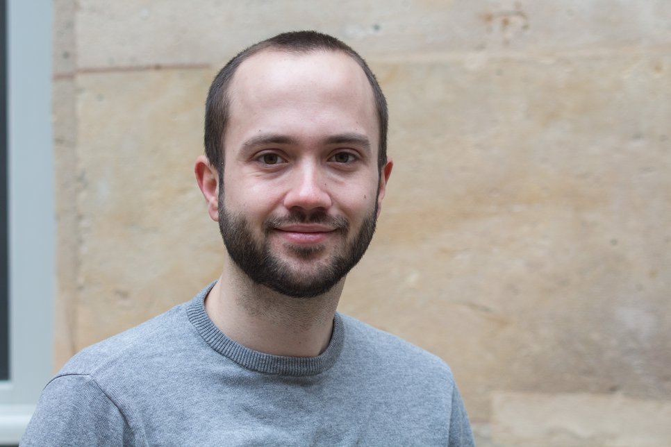 A smiling man with a beard wearing a gray sweater stands in front of a stone wall.