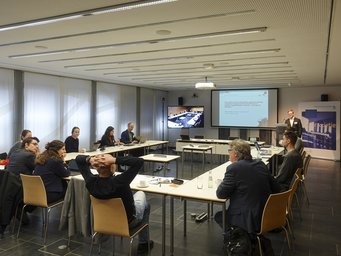 A group of people engaged in a meeting around a conference table in a modern room, with a presentation displayed on a screen and an additional screen showing a remote meeting.