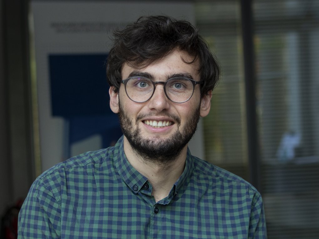 A young man with glasses and curly hair is smiling, wearing a green checkered shirt, in a well-lit indoor setting.