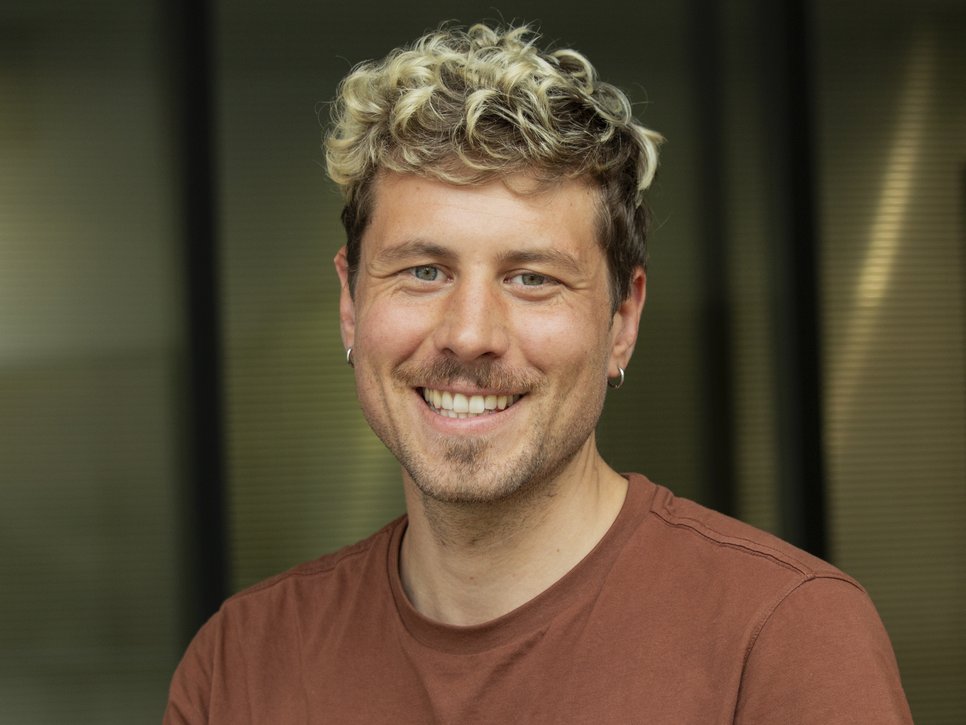 A smiling man with curly blonde hair, wearing a brown shirt, against a blurred background.