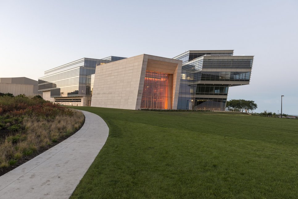 A modern building with glass and stone elements, surrounded by green grass and a pathway. The building features large windows and geometric shapes, suggesting a contemporary architectural design.