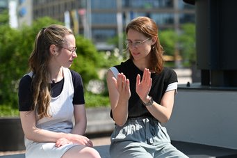 Two women are sitting outdoors, engaged in conversation. One has long, wavy hair and glasses, wearing a black top with sleeves. The other has straight hair, slightly wavy and also wears glasses, dressed in a black top with light-colored sleeves. Green foliage and a city background are visible.