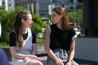 Two women are sitting outdoors, engaged in conversation. One has long, wavy hair and glasses, wearing a black top with sleeves. The other has straight hair, slightly wavy and also wears glasses, dressed in a black top with light-colored sleeves. Green foliage and a city background are visible.