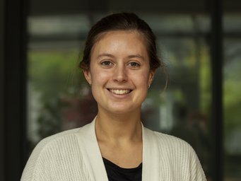 Woman with brown hair tied back, wearing a white knitted cardigan over a black shirt, standing indoors near a large window with blurred greenery outside.