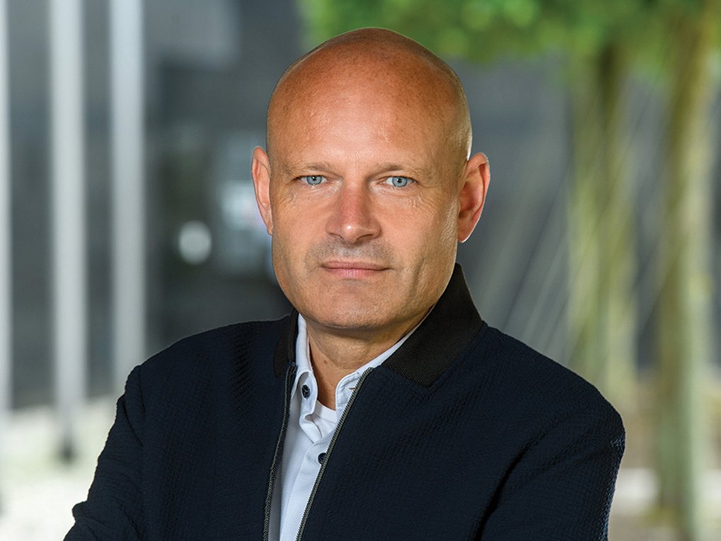A man with a shaved head and blue eyes is looking at the camera, wearing a black jacket over a white shirt, with a blurred background that includes greenery.
