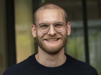 A smiling man with glasses and a beard, wearing a black sweater, in front of a blurred background.