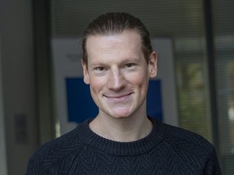 A smiling man with long hair, wearing a dark sweater, is posing for a portrait against a blurred background.