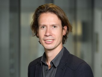 A professional portrait of a man with medium-length, wavy brown hair, wearing a dark blazer and shirt, smiling at the camera.