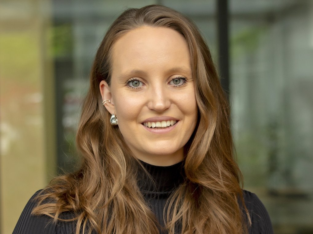 A smiling woman with long, wavy brown hair wearing a black turtleneck and earrings, standing in front of a blurred background.