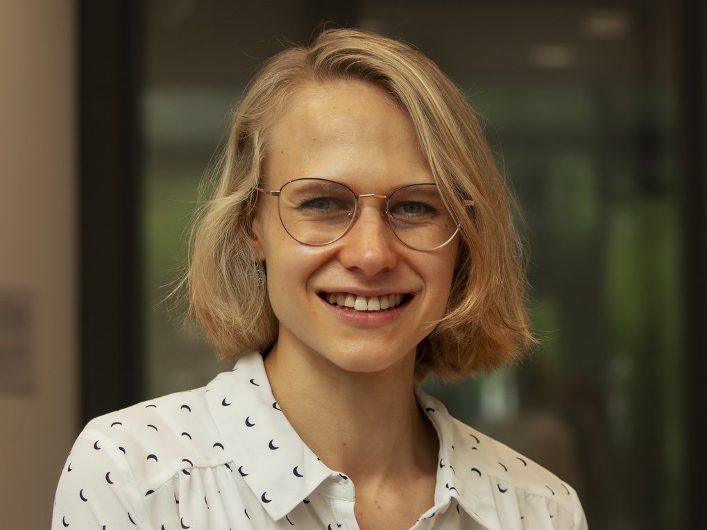 A young woman with short blonde hair and glasses is smiling while wearing a patterned shirt in an indoor setting.