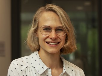 A young woman with short blonde hair and glasses is smiling while wearing a patterned shirt in an indoor setting.