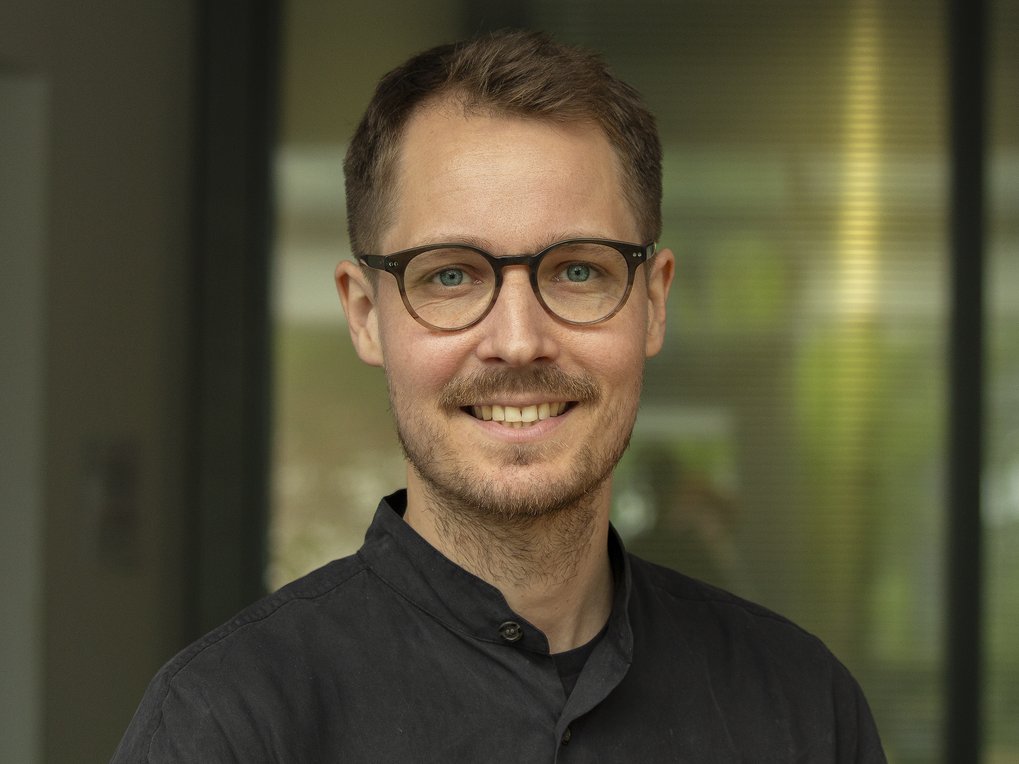 A smiling man with glasses, wearing a dark shirt, standing in front of a blurred background.