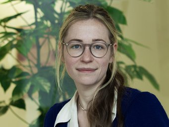 A woman with glasses and long hair, wearing a blue sweater over a white shirt, poses in front of a green plant background.