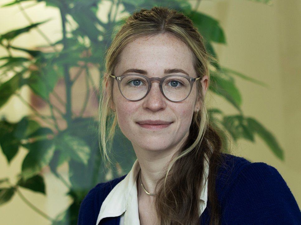 A woman with glasses and long hair, wearing a blue sweater over a white shirt, poses in front of a green plant background.