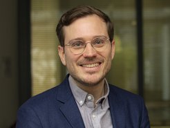 A smiling man with glasses is wearing a blue blazer and a light shirt. The background features blurred indoor elements.