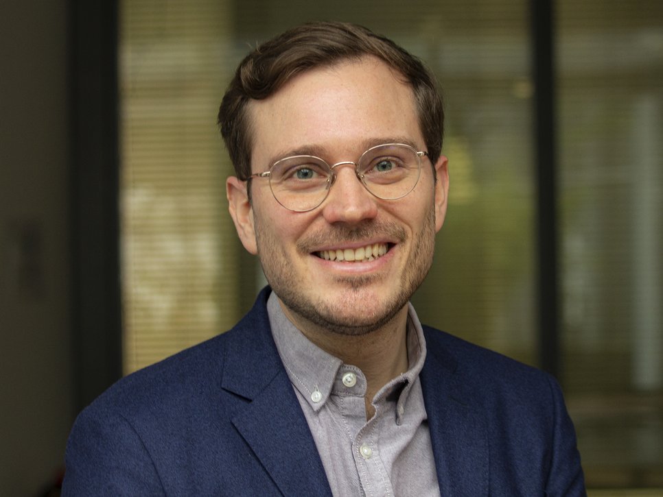 A smiling man with glasses is wearing a blue blazer and a light shirt. The background features blurred indoor elements.