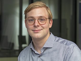 A young man with glasses smiles for the camera, wearing a light blue shirt, in a modern indoor setting.