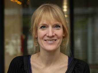 A smiling woman with blonde hair and earrings, wearing a black top, stands in front of a blurred background with soft lighting.