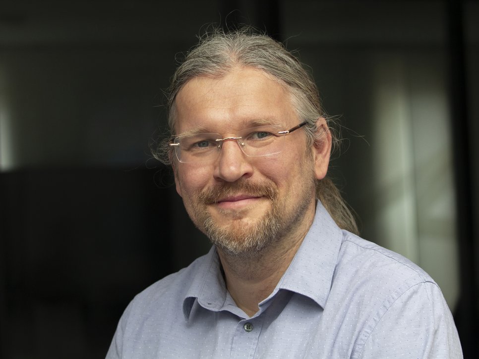 A smiling man with long hair and glasses, wearing a light blue shirt.